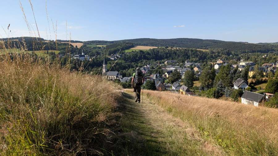 Wanderer auf einem Grasweg im Abstieg, darunter ein Ort mit Kirche